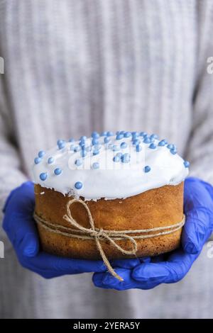 Die Hände des Kochs in blauen Handschuhen halten den Osterkuchen mit weißem Belag und blauen Streuseln. Frau mit traditionellem russischen Osterkuchen. Hausgemachter Kuchen Stockfoto