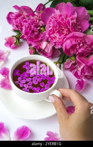 Kreatives Layout mit rosa Pfingstrosen-Blumen. Weibliche Hand hält eine Tasse Tee auf einem hellen Tisch. Frühling saisonal valentine, Frau, Mutter, 8 märz Urlaub, Stockfoto