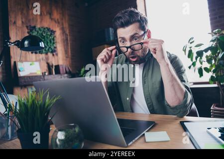 Foto von gutaussehendem jungen Mann erstaunter Blick Netbook ablegen Brille gekleidet grünes Hemd komfortables Startup Büro Loft Raum Innenraum Stockfoto