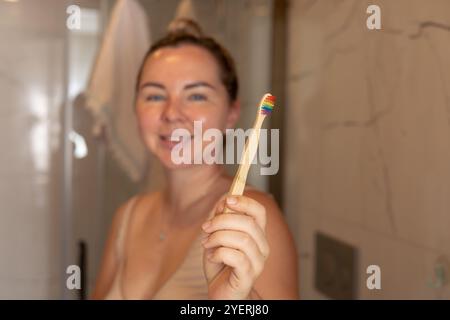 Die lächelnde junge Frau zeigt eine Zahnbürste mit Regenbogenstreifen, während sie in einem gut beleuchteten Badezimmer steht. Sie scheint ihre morgendliche Routine zu genießen und zu fördern Stockfoto