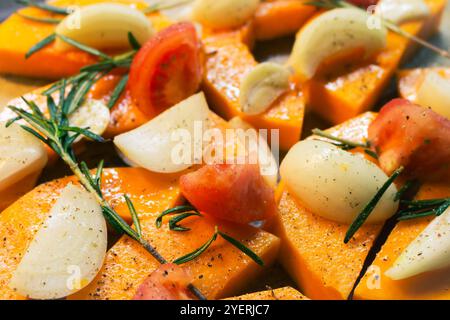 Kürbis in Scheiben mit Zwiebeln, Knoblauch und Rosmarinblättern auf Backblech. Herbstessen Stillleben. Rohe Kürbisscheiben mit Kräutern und Gewürzen, Nahaufnahme. Stockfoto