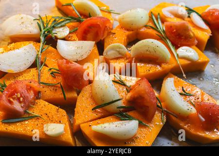 Kürbis in Scheiben mit Zwiebeln, Knoblauch und Rosmarinblättern auf Backblech. Herbstessen Stillleben. Rohe Kürbisscheiben mit Kräutern und Gewürzen, Nahaufnahme. Stockfoto