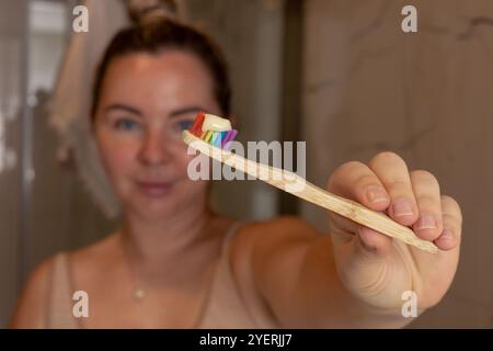 Die lächelnde junge Frau zeigt eine Zahnbürste mit Regenbogenstreifen, während sie in einem gut beleuchteten Badezimmer steht. Sie scheint ihre morgendliche Routine zu genießen und zu fördern Stockfoto