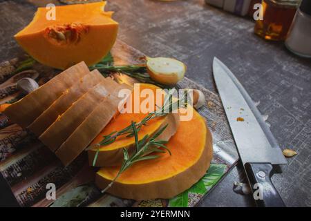 Kürbis in Scheiben mit Zwiebeln, Knoblauch und Rosmarinblättern auf Schneidebrett. Herbstessen Stillleben. Rohe Kürbisscheiben mit Kräutern und Gewürzen und Messer Stockfoto