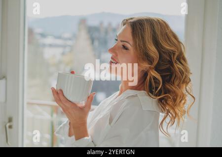 Porträt einer schönen jungen Frau, 42 Jahre alt, mit einer Tasse heißem Tee am Fenster. Gemütliches Zuhause. Stockfoto