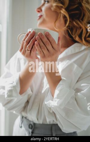 Porträt einer schönen jungen Frau, 42 Jahre alt, mit einer Tasse heißem Tee am Fenster. Gemütliches Zuhause. Stockfoto