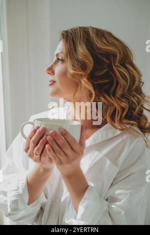 Porträt einer schönen jungen Frau, 42 Jahre alt, mit einer Tasse heißem Tee am Fenster. Gemütliches Zuhause. Stockfoto