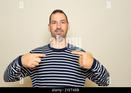 Bärtiger hispanischer Mann in seinen 40ern, der einen gestreiften Pullover trägt und auf seine Arbeit stolz zeigt. Stockfoto