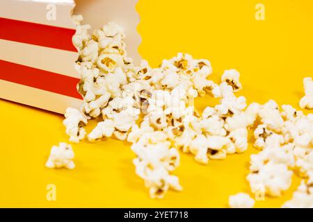 Nahaufnahme eines umgefallenen Kartons mit verstreutem Popcorn auf gelbem Hintergrund, Kinolebensmittelkonzept Stockfoto