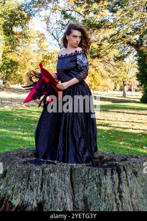 Eine Hexe in einem langen schwarzen Kleid und einem Hexenhut sitzt im Wald des Parks, geschützt vor der rauen Herbstsonne, Schottland Stockfoto