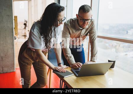 Ein junger zufriedener Mann spricht mit einer Kollegin und freut sich über die geleistete Arbeit. Kollegen arbeiten im Büro an einem Laptop. Arbeiten Sie in einem modernen Büro Stockfoto