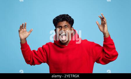 Porträt eines emotionalen Indianers in rotem Hoodie, der die Hände nach oben hebt und mit schockierten und aufgeregten Emotionen vor blauem Studiohintergrund brüllt Stockfoto