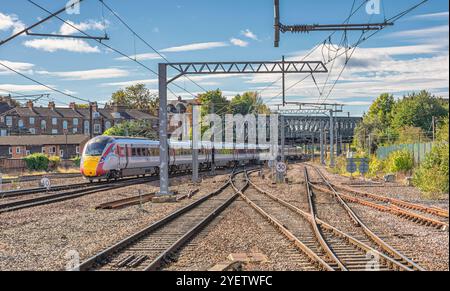 Ein Zug fährt von einem Bahnhof ab und unterquert eine eiserne Brücke. Eisenbahngleise treffen aufeinander und im Hintergrund befinden sich Häuser. Stockfoto