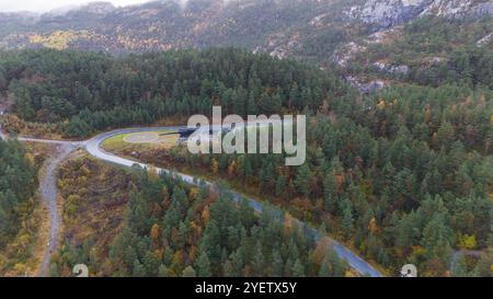 Aus der Vogelperspektive auf eine gewundene Straße durch einen dichten Wald mit Herbstlaub, umgeben von felsigen Hügeln. Ein Hubschrauberlandeplatz ist in der Nähe der Straße und des Landes sichtbar Stockfoto