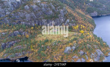 Blick aus der Vogelperspektive auf eine abgelegene Hütte, umgeben von Herbstlaub und felsigen Klippen in der Nähe eines Sees. Stockfoto