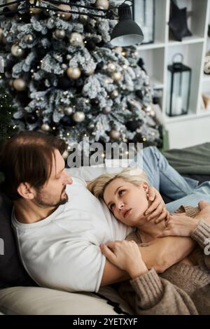 Ein Paar entspannt sich zu Hause, umgeben von einem wunderschön dekorierten Weihnachtsbaum und sanfter Beleuchtung. Stockfoto