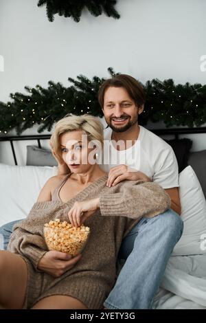 Ein Paar sitzt zusammen auf dem Bett, teilt Popcorn und lächelt in gemütlicher Atmosphäre. Stockfoto