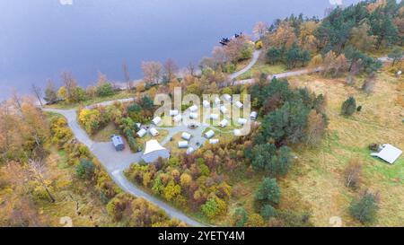 Blick aus der Vogelperspektive auf einen Campingplatz am See mit mehreren Zelten, die in einem Kreis angeordnet sind, umgeben von Herbstbäumen und einer gewundenen Straße. Der See ist auf dem sichtbar Stockfoto