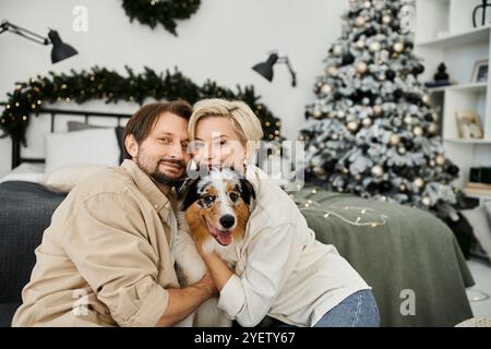 Ein fröhliches Paar kuschelt mit ihrem Hund in einem wunderschön eingerichteten Wohnzimmer für die Feiertage. Stockfoto