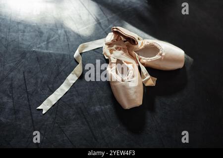 Hintergrundaufnahme von beigefarbenen Satin spitzenschuhen mit eleganten Bändern, die im Sonnenlicht auf dunklem Boden im Tanzstudio liegen, Kopierraum Stockfoto