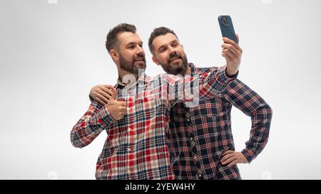 Zwei junge Männer, Zwillingsbrüder kleideten passende karierte Hemden und Jeans, Umarmungen und Selfie vor weißem Studiohintergrund. Stockfoto