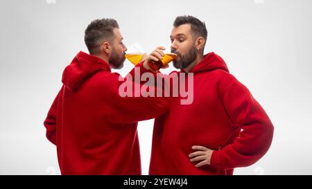 Zwei gleich gekleidete junge Männer, Zwillingsbrüder, trinken Craft-Bier aus Glasbechern vor weißem Studiohintergrund. Stockfoto