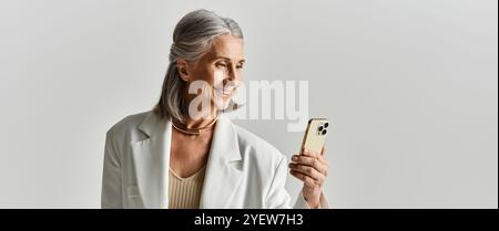 Eine anmutige reife Frau in einem weißen Blazer genießt einen Moment der Freude, während sie in einer schicken Umgebung an ihrem Handy lächelt. Stockfoto