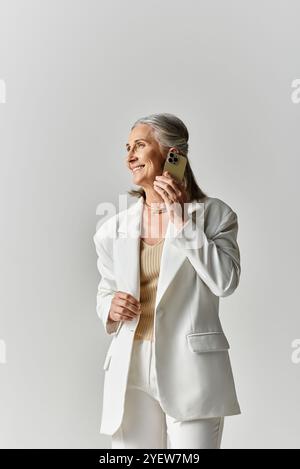 Eine reife Frau strahlt Charme und Anmut aus und lächelt in einem schicken weißen Anzug vor grauer Kulisse. Stockfoto