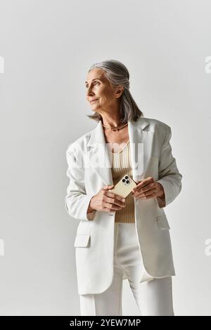 Eine reife Frau mit schönen grauen Haaren steht selbstbewusst in einem maßgeschneiderten weißen Anzug und hält ihr Handy. Stockfoto