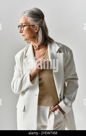 Eine raffinierte Frau verströmt Selbstvertrauen in einem weißen Blazer und einem beigefarbenen Oberteil vor grauem Hintergrund. Stockfoto