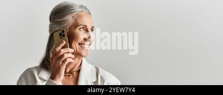 Eine stilvolle, reife Frau strahlt Selbstvertrauen aus, während sie anmutig mit ihrem Telefon spricht, in weiß gekleidet. Stockfoto