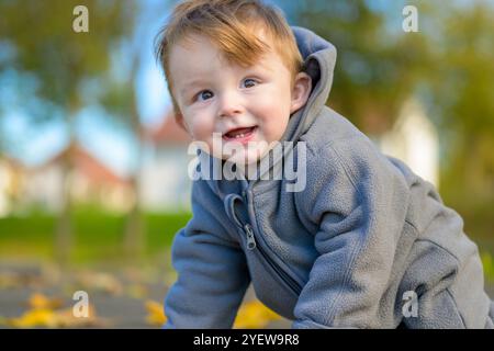 Ein fröhliches Kleinkind in einer gemütlichen grauen Jacke beugt sich nach vorne und blitzt ein helles Lächeln auf, das die Szene erleuchtet. Stockfoto