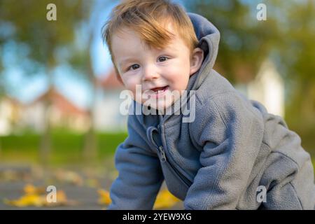 Ein fröhliches Kleinkind in einer warmen grauen Jacke lächelt hell, während es die Natur erkundet. Stockfoto