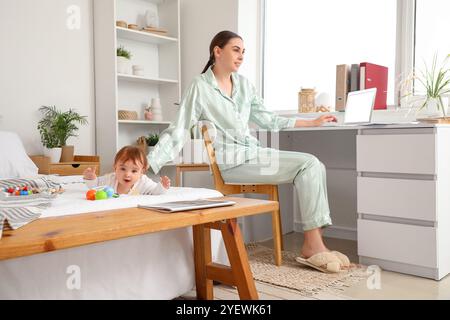 Junge Geschäftsmutter im Pyjama, die mit Laptop arbeitet und kleines Baby, das zu Hause spielt Stockfoto