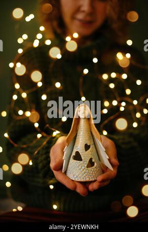 Nahaufnahme der Hände, die Engelsfigur mit weihnachtlichem Bokeh-Hintergrund halten. Frauenhände, die glühende Keramikengel, Concept Christmas und Ange sanft halten Stockfoto