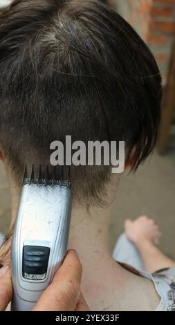 Vertikales Foto eines jungen Mannes, der Haare mit einem elektrischen Trimmer schneidet, Nahaufnahme, Hand mit Haarschneider, der Haare auf dem Hinterkopf des Teenagers schneidet Stockfoto