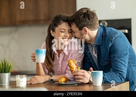 Ein freudiges Paar teilt einen warmen Moment beim Frühstück in einer modernen Küche. Sie halten Kaffee und ein Sandwich und strahlen Glück und Liebe aus. Stockfoto