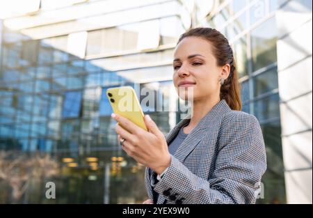 Selbstbewusste Geschäftsfrau in Geschäftskleidung, die auf den Bildschirm ihres Smartphones blickt, während sie draußen vor einem Bürogebäude steht. Stockfoto