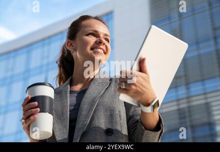 Fröhliche städtische Frau in Business-Freizeitkleidung Stände mit einem thermischen Kaffeetasse und einem digitalen Tablet in der Hand in der Stadt und lächelnd. Stockfoto