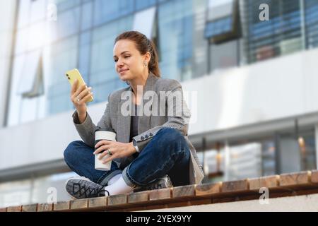 Eine urbane Frau in Business-Freizeitkleidung, die in einer Lotuspose auf einer Bank vor einem Geschäftsgebäude sitzt und ihr Smartphone während ihrer Kaffeepause benutzt Stockfoto