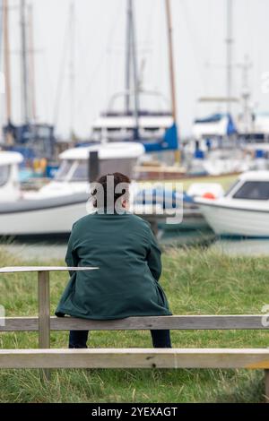 Ältere oder ältere Dame mittleren Alters, die allein auf einer Holzbank am Meer sitzt und einen geschäftigen Yachthafen in Yarmouth auf der isle of wight, Großbritannien, überblickt Stockfoto