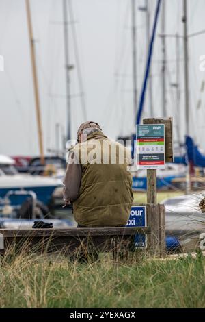 Mann, der allein an der Seite eines Yachthafens oder Yachthafens sitzt, trägt eine flache Kappe mit Segelyachten und Booten im Hintergrund. Stockfoto