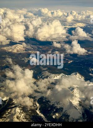 Blauer Himmel mit Wolken vor, hinter, über und unter werden von einem Flugzeugfenster aus gesehen. Dies ist ein dramatischer Blick auf die Berge unter Ihnen. Siehe Schnee unten. Stockfoto