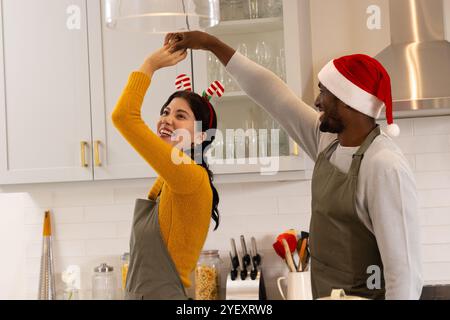 Tanzende, multirassische Paare mit festlichen Hüten und Schürzen, die weihnachten feiern, zu Hause Stockfoto