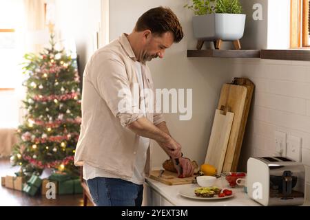 Senior Mann, der festliche Mahlzeit in der Küche mit Weihnachtsbaum im Hintergrund zubereitet, zu Hause Stockfoto