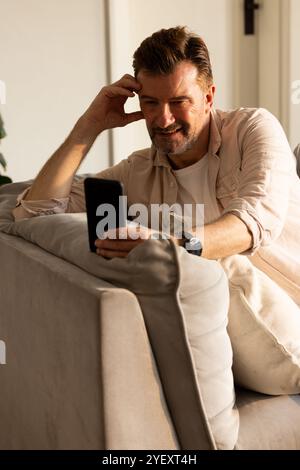 Lächelnder älterer Mann, der sich während der Ferien mit dem Smartphone auf der Couch entspannt Stockfoto