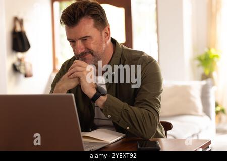 Reifer Senior Mann lächelt, während er zu Hause mit dem Laptop unterwegs ist und gerne Online-Weihnachtseinkäufe genießt Stockfoto