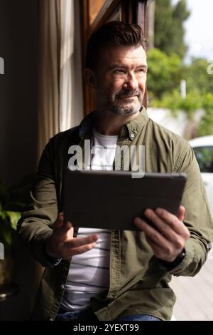 Seniorenmann, der ein Tablet am Fenster benutzt und zu Hause einen friedlichen Moment genießt Stockfoto