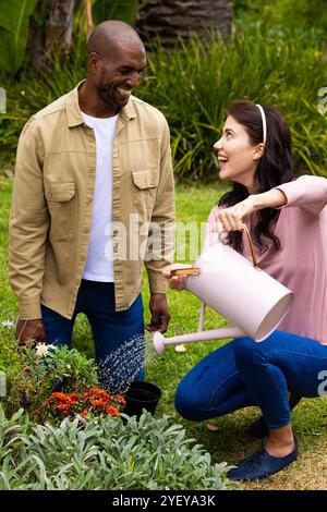 Ein multirassisches Paar, das zusammen im Garten gärtet, lächelt und Pflanzen im Hinterhof tränkt Stockfoto