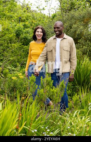 Multirassisches Paar, das Hand in Hand durch den üppigen grünen Garten geht und glücklich lächelt Stockfoto